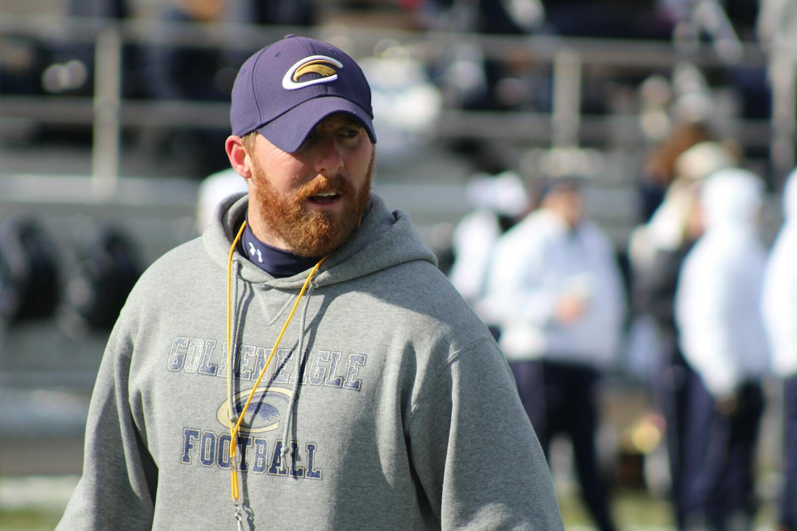 A focused football coach in a hoodie and cap on the sidelines of a stadium, ready to lead the team.